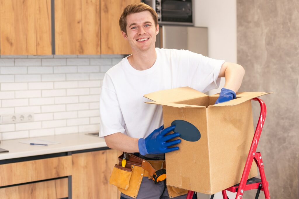 Smiling male worker of moving and delivery company holding cardboard box. Loader in overalls posing